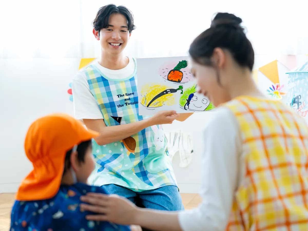 イラストを持つ笑顔の男性保育士と子供と接する女性保育士の画像