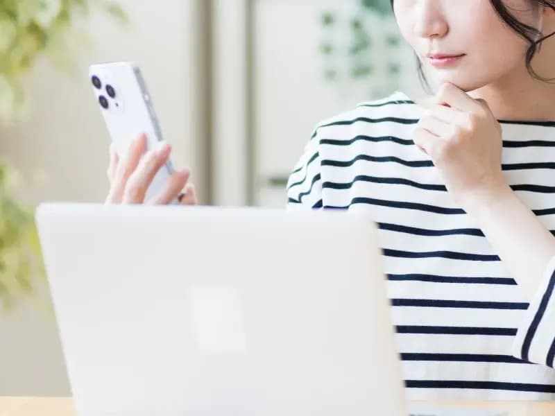 ノートパソコンを開きスマホを見て考える女性のイメージ