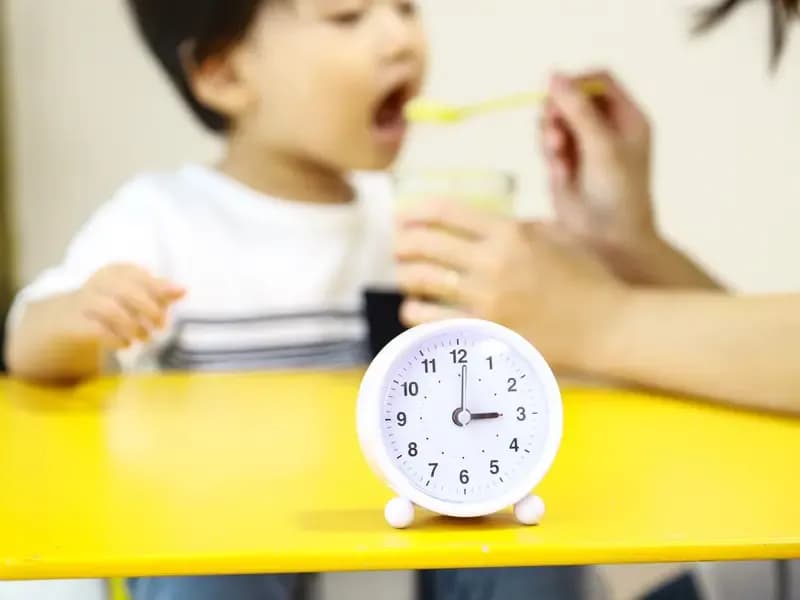 時計とおやつを食べる園児と介助する保育士のイメージ