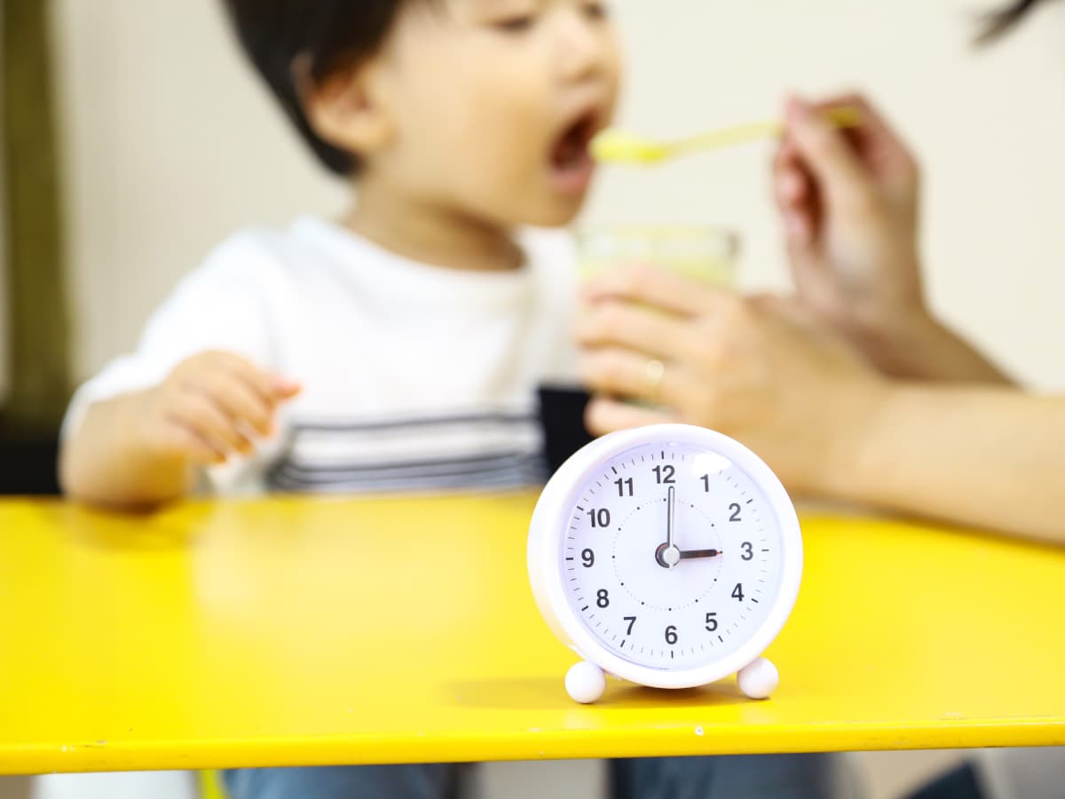 時計とおやつを食べる園児と介助する保育士の画像