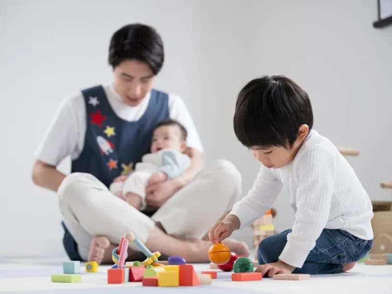 赤ちゃんをあやす保育士と一人で遊ぶ子供のイメージ
