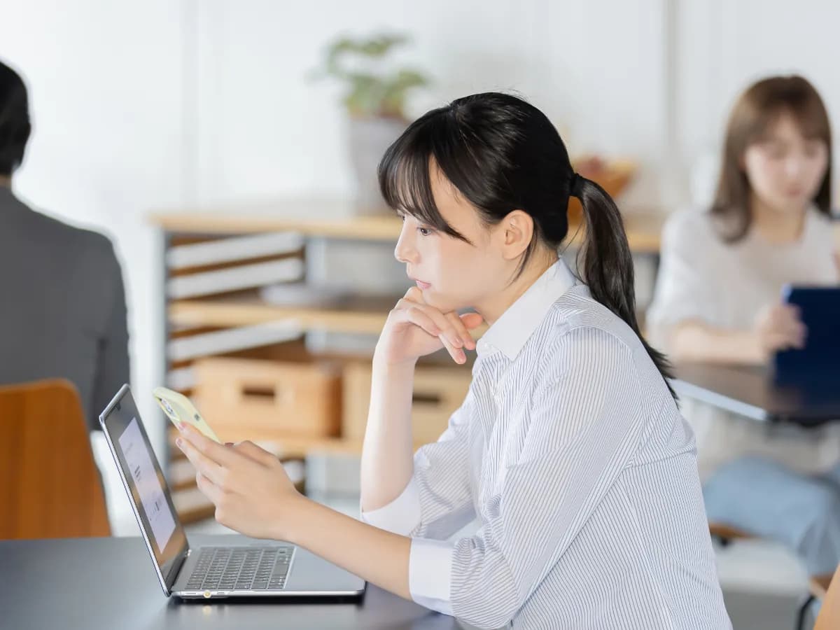 ノートパソコンを広げながらスマートフォンを眺める若い女性の画像
