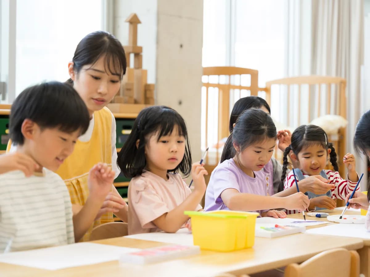 絵の具を使って絵を描いている子供たちと見守っている女性児童指導員のイメージ