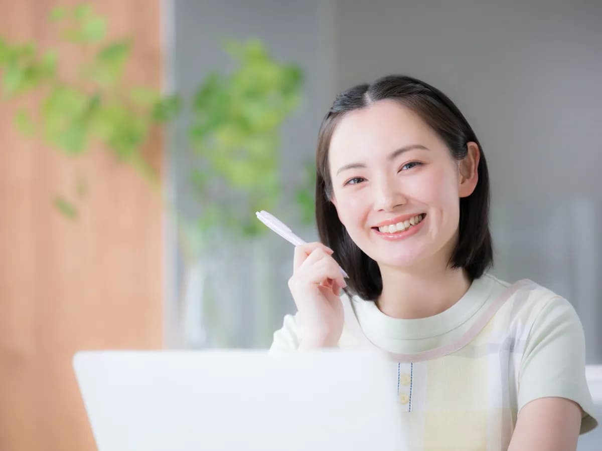 ノートパソコンを開きペンを持ってほほ笑む女性保育士のイメージ
