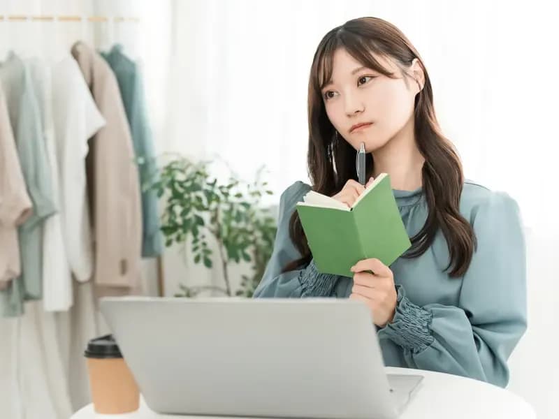 パソコンの前でスケジュール帳を持ちながら悩む若い女性のイメージ