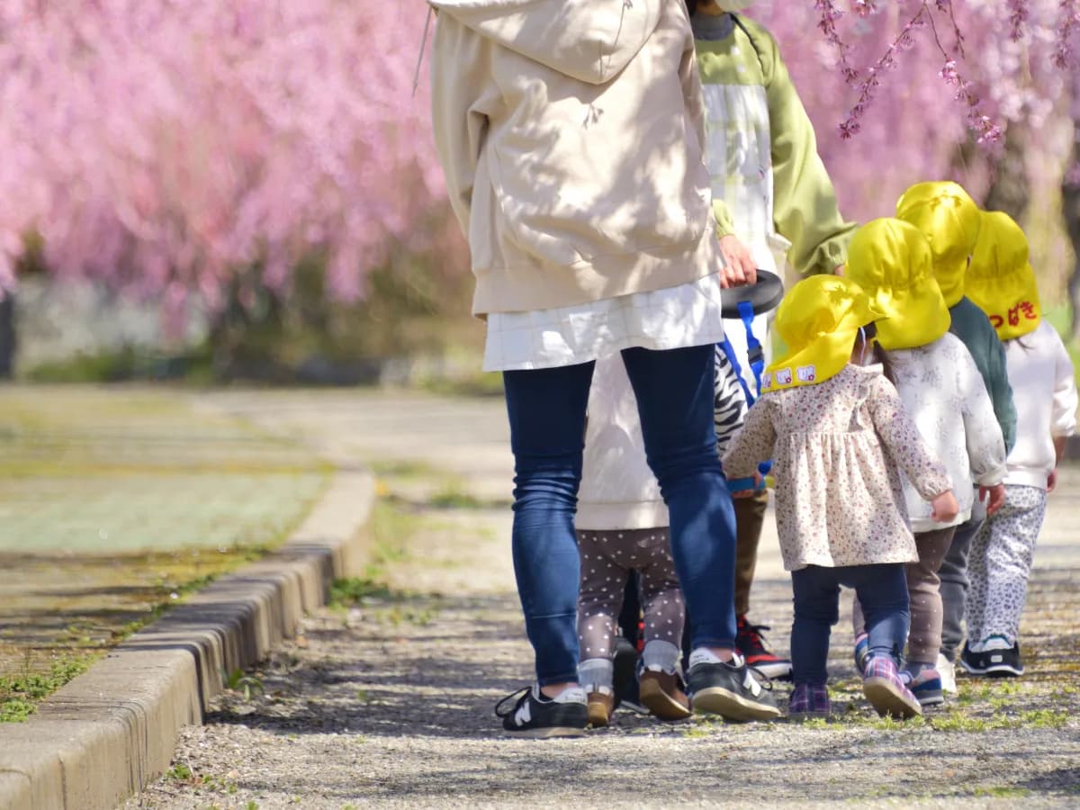 春の公園を散歩する保育士と保育園児達の画像
