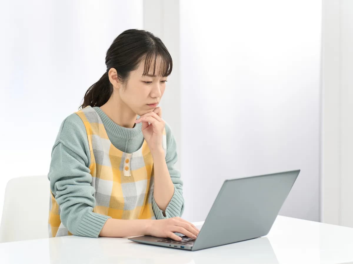 顎に手を当て真剣な表情でノートパソコンを見ている女性の保育士の画像